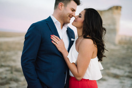 A couple stands close together outdoors, smiling and about to kiss. The man wears a blue suit and white shirt; the woman has long dark hair and wears a white top and red skirt. The background is a blurred, sandy landscape. by Justin Salem Meyer
