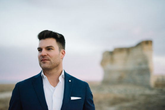 A man in a dark blue suit and white shirt stands outdoors with a blurred rock formation in the background under a cloudy sky. by Justin Salem Meyer