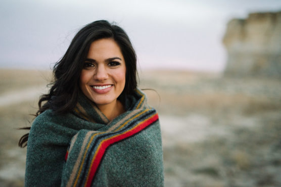 A woman with long dark hair smiles at the camera, wrapped in a gray blanket with red and yellow stripes, standing outdoors in a blurred, open landscape. by Justin Salem Meyer