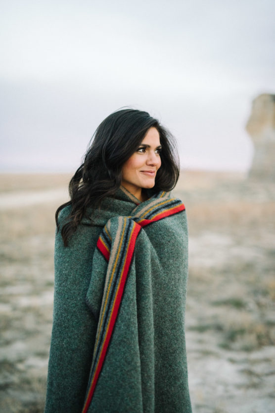 A woman with long dark hair stands outdoors, wrapped in a thick green blanket with red and yellow stripes, looking off to the side. The background shows a blurred, rocky landscape under a cloudy sky. by Justin Salem Meyer