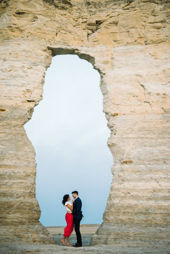 A couple stands facing each other and embracing beneath a large, natural rock archway, with pale stone cliffs surrounding them and a soft blue sky in the background. by Justin Salem Meyer