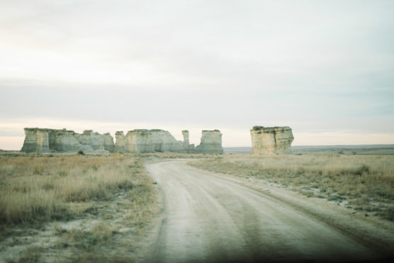 A dirt road curves through dry grassland toward large, flat-topped rock formations under a pale, cloudy sky, creating a serene, open landscape. by Justin Salem Meyer