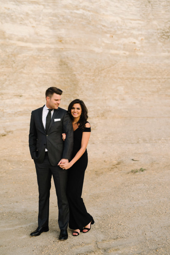 A man in a dark suit and a woman in a long black dress stand close together on a sandy, beige landscape. She smiles at the camera while holding his arm; he looks off to the side. by Justin Salem Meyer