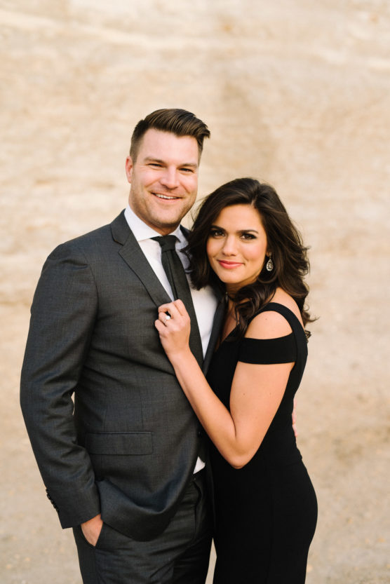 A man in a dark suit and tie stands next to a woman in a black, off-the-shoulder dress. They are smiling and embracing each other while posing outdoors against a light, sandy background. by Justin Salem Meyer