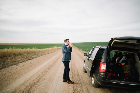 A man in a suit stands on a rural dirt road beside an open car trunk filled with bags, looking into the distance. Fields and open sky stretch out in the background. by Justin Salem Meyer
