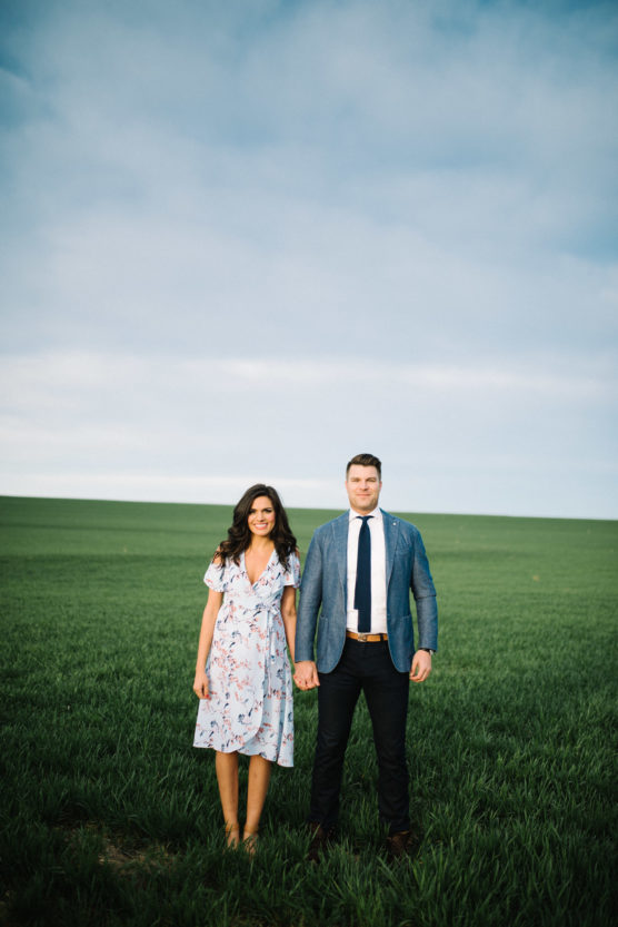 A woman in a light floral dress and a man in a blue blazer with a tie stand holding hands in a green field under a blue sky with light clouds. by Justin Salem Meyer
