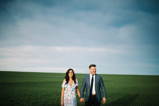 A woman in a floral dress and a man in a suit hold hands, smiling as they walk through a wide green field under a partly cloudy blue sky. by Justin Salem Meyer