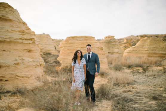 A woman in a floral dress and a man in a suit stand smiling among beige rock formations in a dry, grassy landscape under a cloudy sky. by Justin Salem Meyer