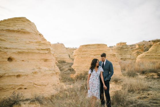 A couple walks together among yellow sandstone rock formations and dry grass on a cloudy day. The woman wears a light floral dress, and the man is dressed in a suit. They appear happy and relaxed outdoors. by Justin Salem Meyer