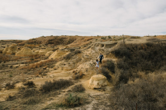 Two people walk along a dirt trail through a dry, rocky landscape with low hills and sparse vegetation under a cloudy sky. by Justin Salem Meyer