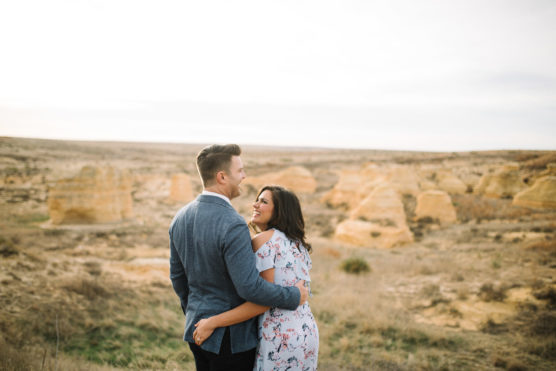 A couple stands outdoors, embracing and smiling at each other, with a scenic backdrop of rocky, desert-like formations and dry grass under a bright, cloudy sky. by Justin Salem Meyer