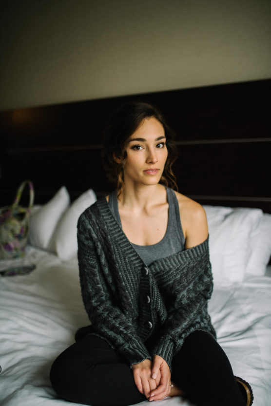 A young woman with wavy brown hair sits on a bed, wearing a gray tank top and a chunky knit cardigan that falls off one shoulder. She looks calmly at the camera, hands loosely clasped in her lap. Pillows and a bag are in the background. by Justin Salem Meyer