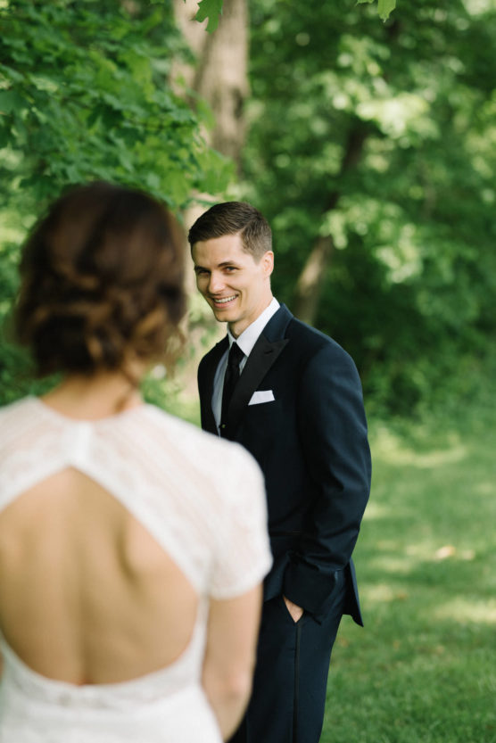 A groom in a black suit smiles warmly at his bride, who stands with her back to the camera in a white dress with an open back. They are outdoors, surrounded by lush green trees. by Justin Salem Meyer