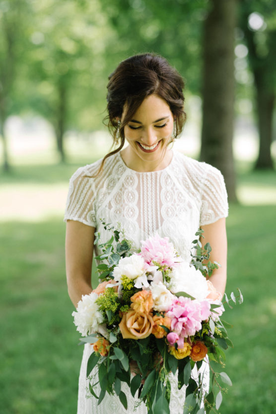 A woman in a white lace dress stands outdoors, smiling as she looks down at a large, colorful bouquet of flowers. The background is green and leafy, with trees and soft sunlight. by Justin Salem Meyer