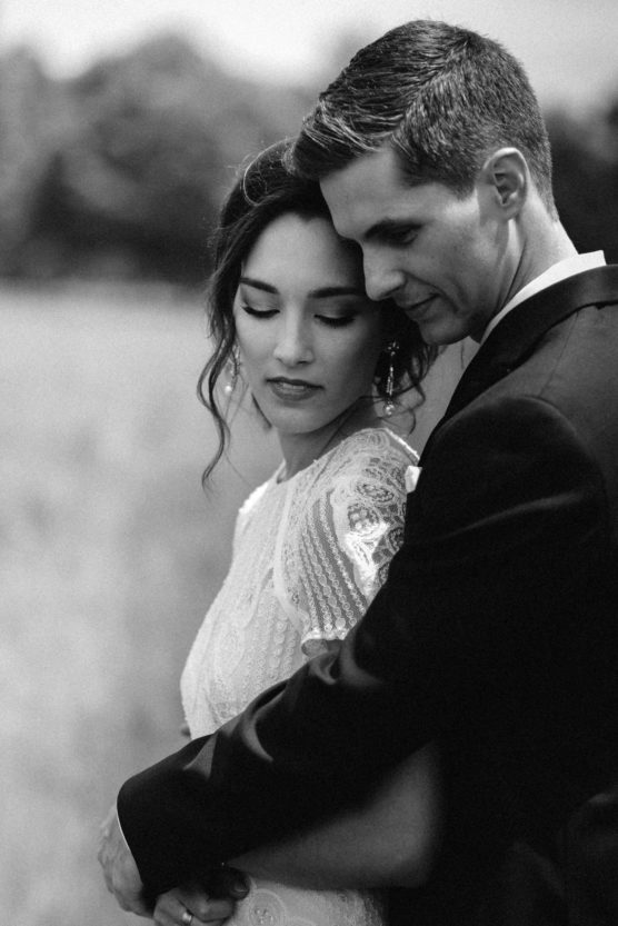 A black and white photo of a couple in formal attire standing close together outdoors. The woman, in a lacy dress, looks down with a gentle expression while the man, in a suit, embraces her from behind. by Justin Salem Meyer