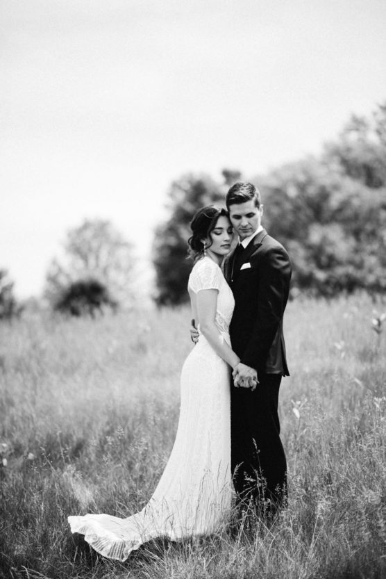 A bride and groom stand closely together in a grassy field, holding hands and looking serene. The bride wears a white gown, and the groom wears a dark suit. The black-and-white photo has a soft, romantic atmosphere. by Justin Salem Meyer