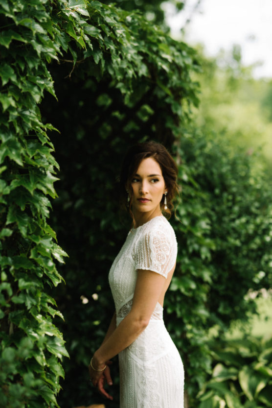 A woman in a lace white dress stands outdoors near lush green foliage, looking slightly over her shoulder. The background is filled with leafy plants and natural greenery. by Justin Salem Meyer