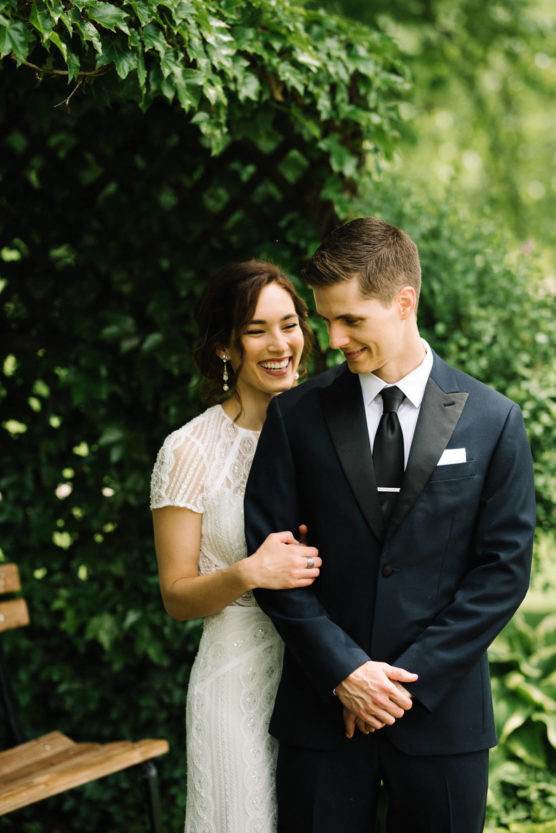 A bride in a white lace dress stands behind a groom in a black suit and tie, both smiling and embracing in a lush green garden setting. by Justin Salem Meyer