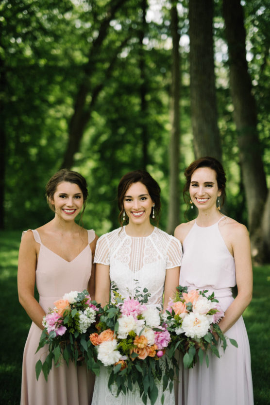 Three women stand outdoors in formal dresses, holding colorful flower bouquets. The woman in the center wears a white lace dress, while the two on either side wear light pink dresses. Trees and greenery fill the background. by Justin Salem Meyer