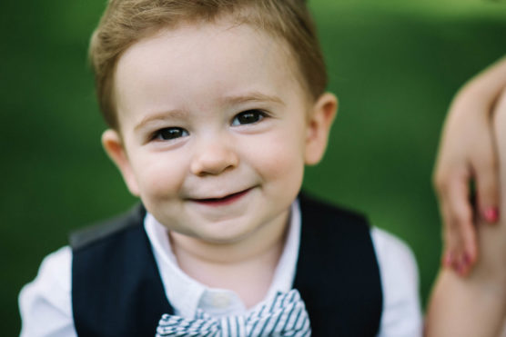 A young boy with light brown hair smiles at the camera, wearing a white shirt, striped bow tie, and dark vest. There is a blurred green background and part of an adult’s hand is visible beside him. by Justin Salem Meyer