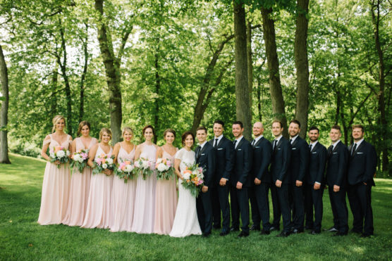 A wedding party stands outdoors on green grass with tall trees in the background. The bride and bridesmaids wear light pink dresses, and the groom and groomsmen wear dark suits. Everyone is smiling and holding bouquets. by Justin Salem Meyer