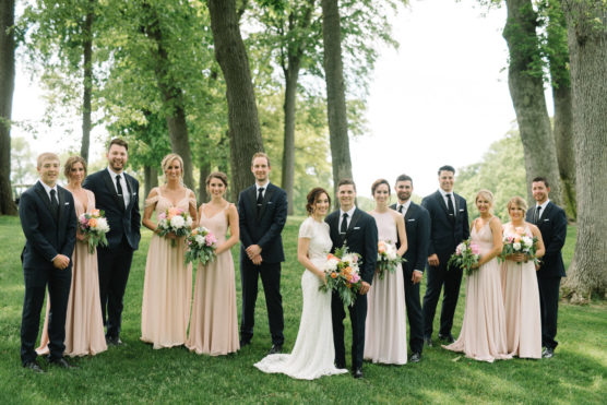 A wedding party poses outdoors among trees. The bride in a white dress and the groom in a black suit stand together, surrounded by bridesmaids in pale pink dresses and groomsmen in black suits, all holding flower bouquets. by Justin Salem Meyer
