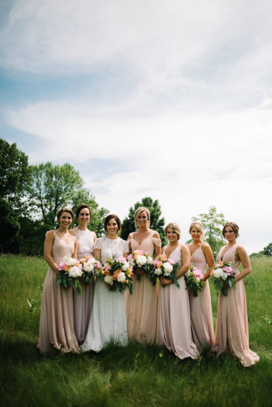A bride in a white dress stands outside with six bridesmaids in blush pink dresses, all holding bouquets of flowers, against a green field and trees under a partly cloudy sky. by Justin Salem Meyer
