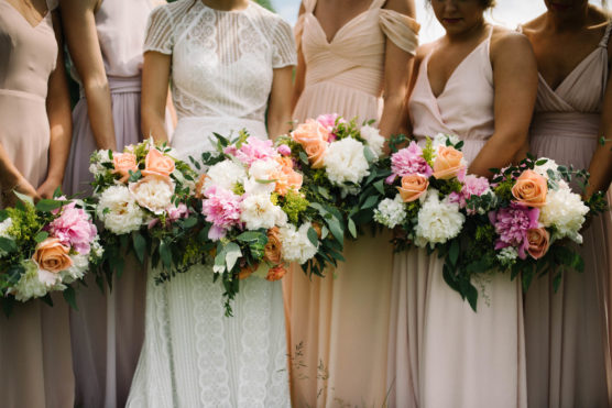 A bride in a white lace dress stands with bridesmaids in blush pink dresses, all holding bouquets of pink, peach, and white flowers with greenery. The photo is cropped to show only their torsos and bouquets. by Justin Salem Meyer