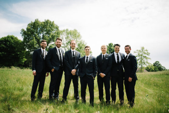 Seven men in black suits and ties stand together, smiling, in a grassy field with green trees and a partly cloudy sky in the background. by Justin Salem Meyer