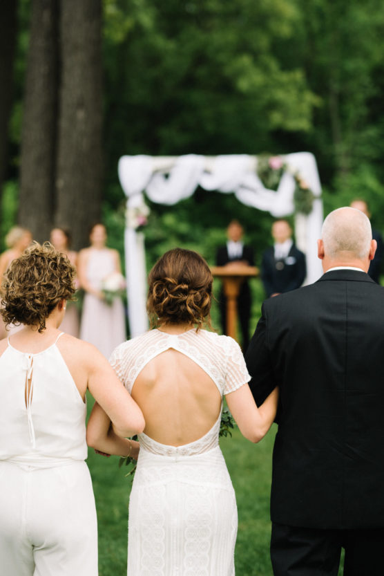 A bride in a white dress with an open back walks arm-in-arm with two people toward an outdoor wedding altar, where guests and an officiant wait. Lush greenery surrounds the ceremony area. by Justin Salem Meyer