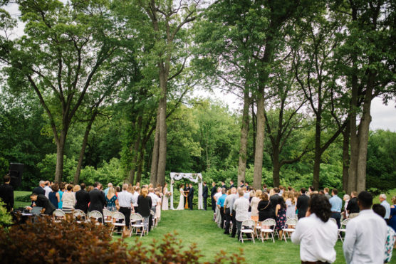 A large group of people attends an outdoor wedding ceremony in a green park, seated in rows facing an altar decorated with white fabric, surrounded by tall trees and lush greenery. by Justin Salem Meyer