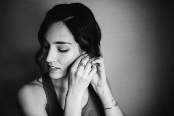A black and white photo of a woman with dark hair, wearing a sleeveless top, gently putting on an earring while looking down. The background is softly blurred. by Justin Salem Meyer