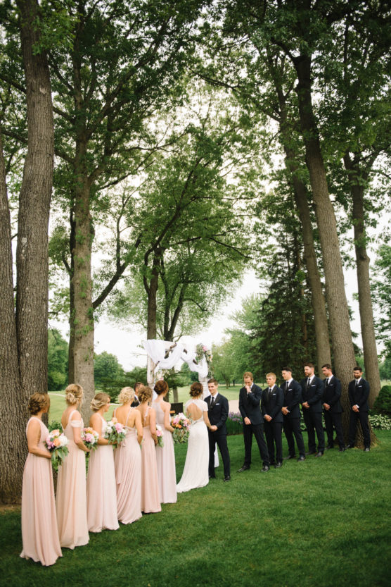 A bride and groom stand facing each other during an outdoor wedding ceremony, surrounded by bridesmaids in light pink dresses and groomsmen in dark suits, beneath tall trees and a decorated arch. by Justin Salem Meyer