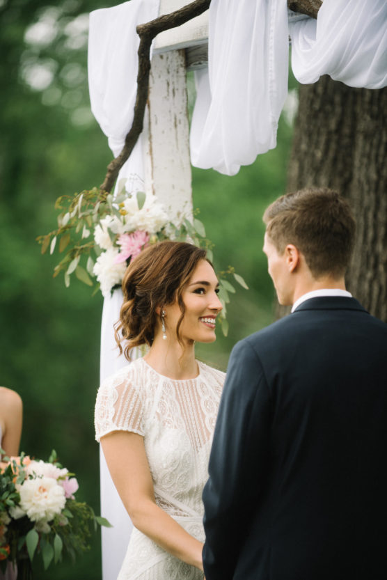 A bride in a white lace dress smiles at the groom during an outdoor wedding ceremony under a decorated wooden arch with white fabric and flowers. Green trees are visible in the background. by Justin Salem Meyer
