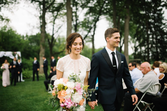 A bride in a white dress holding a colorful bouquet walks hand-in-hand with a groom in a dark suit down an outdoor aisle, smiling, with guests seated on either side and trees in the background. by Justin Salem Meyer