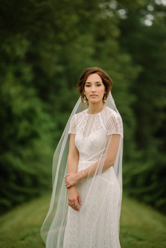 A bride in a lace wedding dress and veil stands outdoors on a grassy path surrounded by lush green trees, looking calmly at the camera with her hands gently clasped. by Justin Salem Meyer