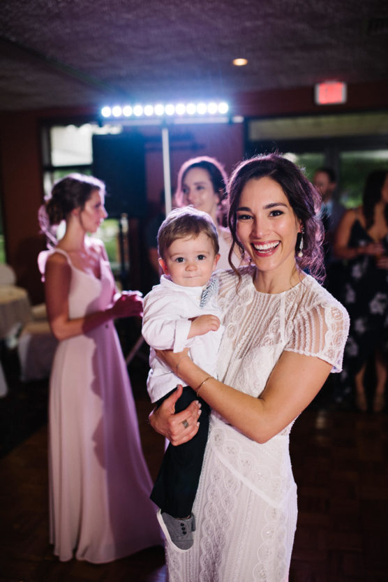 A smiling woman in a white dress holds a baby boy wearing a white shirt at an indoor event, with other people socializing and soft lights in the background. by Justin Salem Meyer