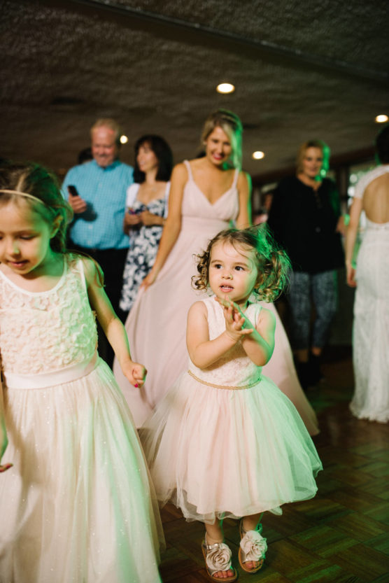 Two young girls in white dresses dance and clap at a wedding reception, with smiling adults and a bride in a white gown watching in the background. The scene is joyful and warmly lit. by Justin Salem Meyer