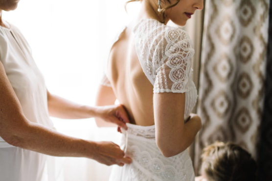 A bride in a white lace dress is being helped with the back of her gown by another person. Soft daylight streams in, highlighting the delicate details of the fabric. The scene feels intimate and calm. by Justin Salem Meyer