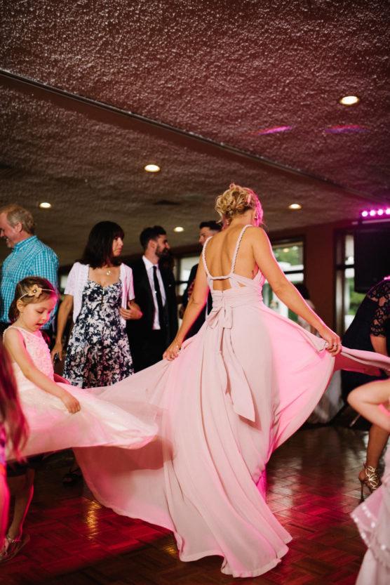 A woman in a long, light pink dress dances indoors, holding her skirt as a young girl in a matching dress plays nearby. Other guests in formal attire are visible in the background. by Justin Salem Meyer