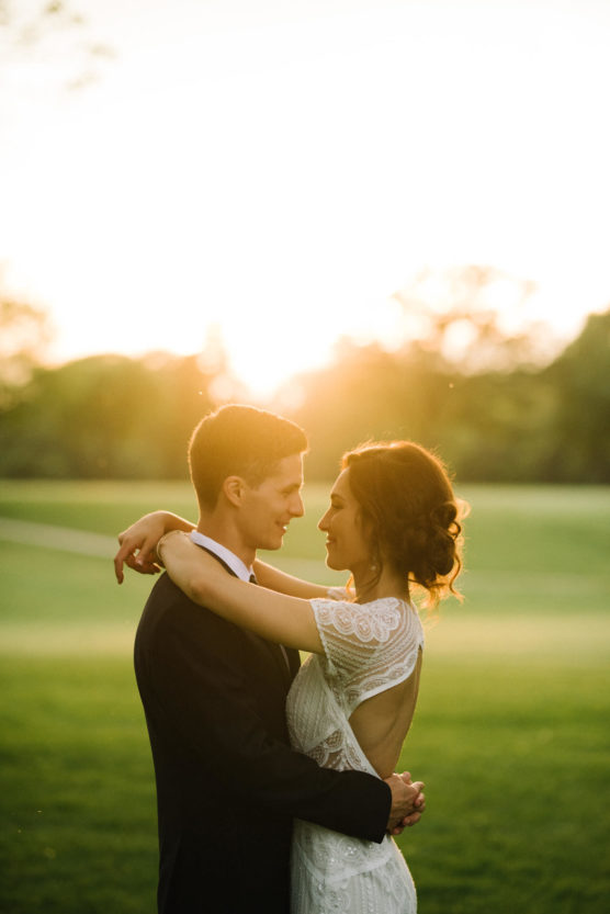 A bride and groom embrace outdoors at sunset, smiling at each other in a green field with soft sunlight glowing behind them. by Justin Salem Meyer