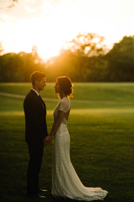 A bride and groom stand facing each other, holding hands on a grassy field at sunset, with warm golden sunlight illuminating the scene and trees in the background. by Justin Salem Meyer