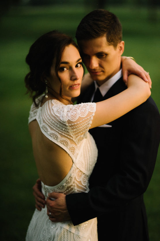 A bride in a white, lace, open-back dress embraces a groom in a black suit outdoors. Both look at the camera with soft expressions, while the background is out of focus and green. by Justin Salem Meyer