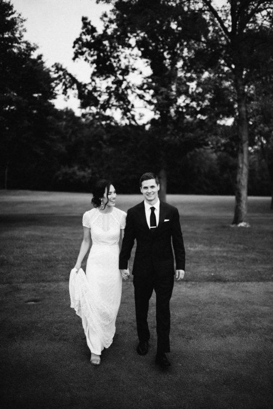 A bride in a white dress and a groom in a black suit walk hand in hand outdoors, smiling at each other, with trees and grass in the background. The photo is in black and white. by Justin Salem Meyer