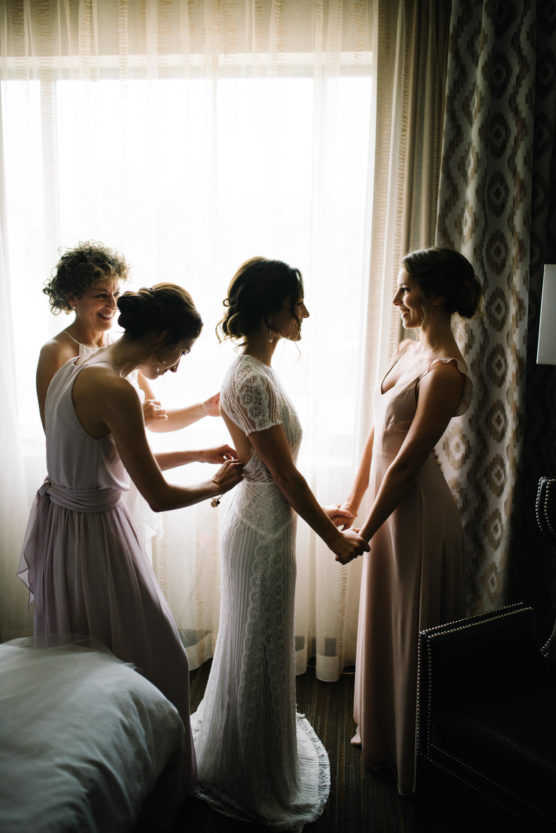Three women help a bride in a white lace dress get ready by fastening her dress in a softly lit room, as she holds hands with one of them and smiles, standing in front of a window with sheer curtains. by Justin Salem Meyer