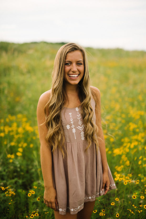 A young woman with long, wavy hair stands smiling in a field of yellow wildflowers, wearing a sleeveless, light brown dress with white embroidery. The background is green and slightly blurred. by Justin Salem Meyer