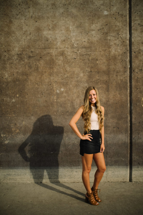 A young woman with long wavy hair stands smiling against a textured concrete wall, wearing a sleeveless white top, black skirt, and brown heeled sandals. Her hand is on her hip and her shadow is visible on the wall. by Justin Salem Meyer