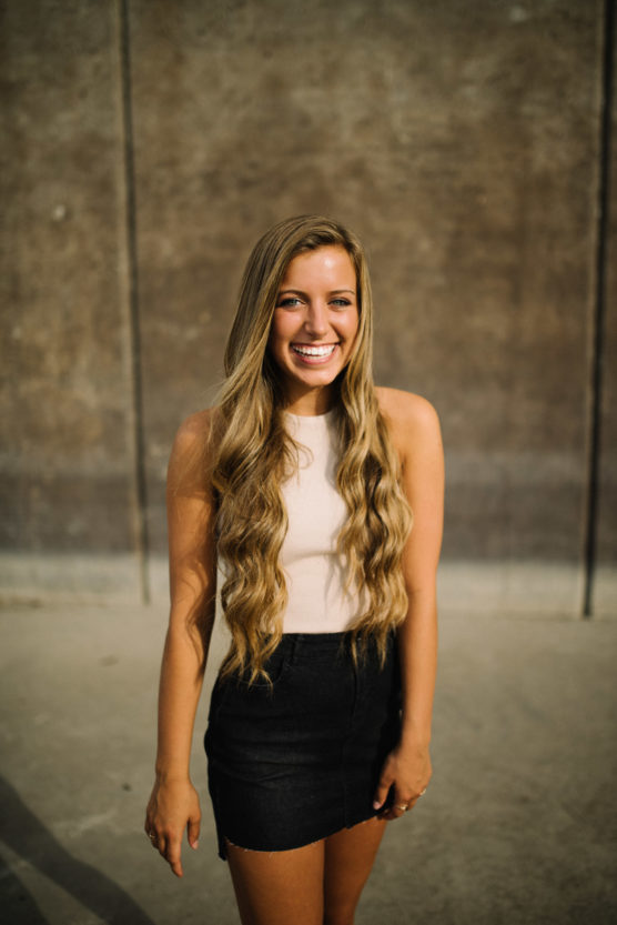 A young woman with long wavy blonde hair, wearing a sleeveless light top and a black skirt, stands outdoors smiling brightly against a textured brown wall. by Justin Salem Meyer