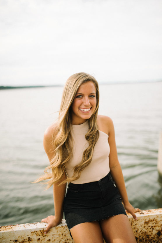 A young woman with long blonde hair, wearing a sleeveless beige top and a black skirt, sits on a ledge by the water, smiling at the camera with a blurred horizon in the background. by Justin Salem Meyer