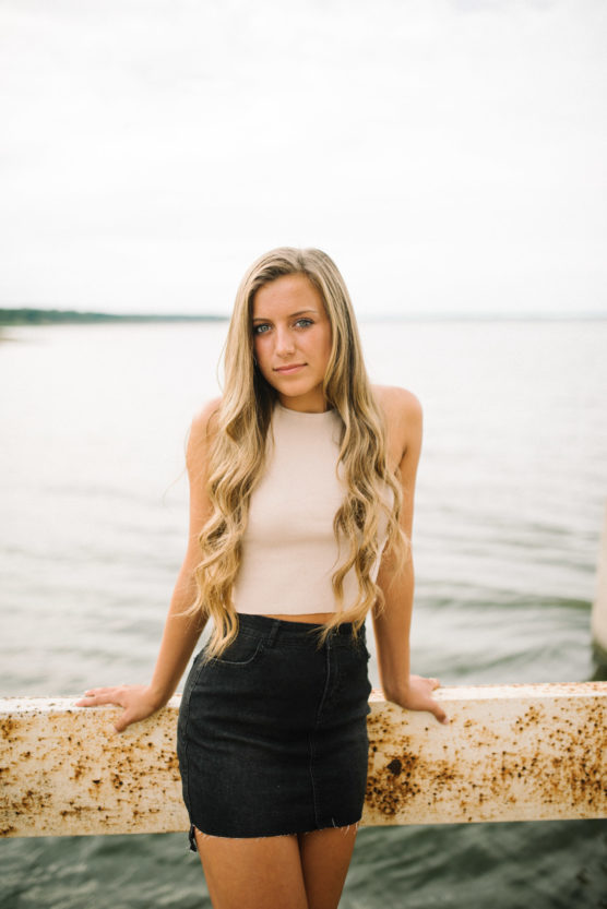 A young woman with long blonde hair, wearing a sleeveless beige top and a black denim skirt, leans against a rusty metal railing by the water on a cloudy day. by Justin Salem Meyer